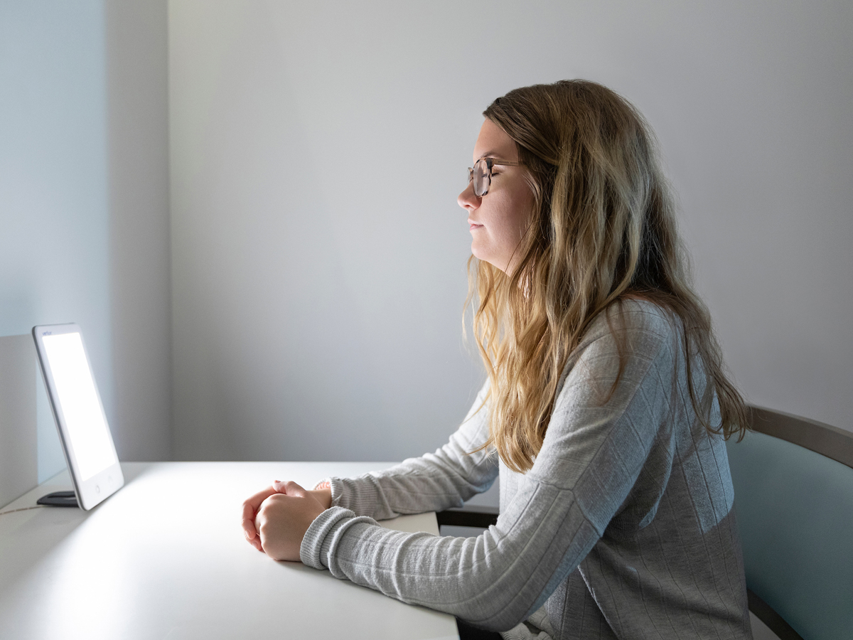 A student sits at a white desk using a bright light therapy lamp in a softly lit room.