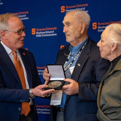 A man presents a medal to an older couple at an academic event with Syracuse University/Newhouse School branding in the background