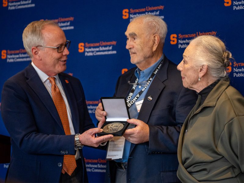A man presents a medal to an older couple at an academic event with Syracuse University/Newhouse School branding in the background