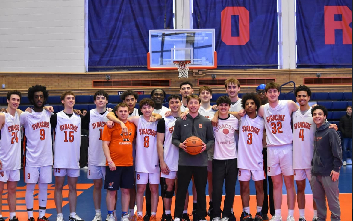 Basketball team in white Syracuse jerseys poses together on an indoor court under the hoop, with blue and orange banners in the background.