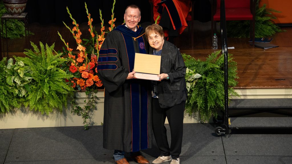 Two people pose together on stage holding an award, one in academic regalia, with orange floral arrangements in the background.