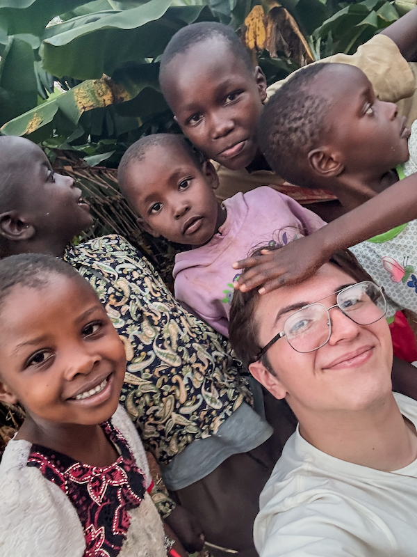 Selfie of a person with glasses surrounded by several children outdoors near leafy plants, with one child’s hand resting on the person’s head. 