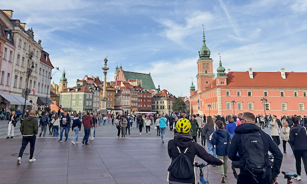 A crowded city square with colorful historic buildings and a tall clock tower, as pedestrians walk and a cyclist wearing a bright yellow helmet passes in the foreground. 