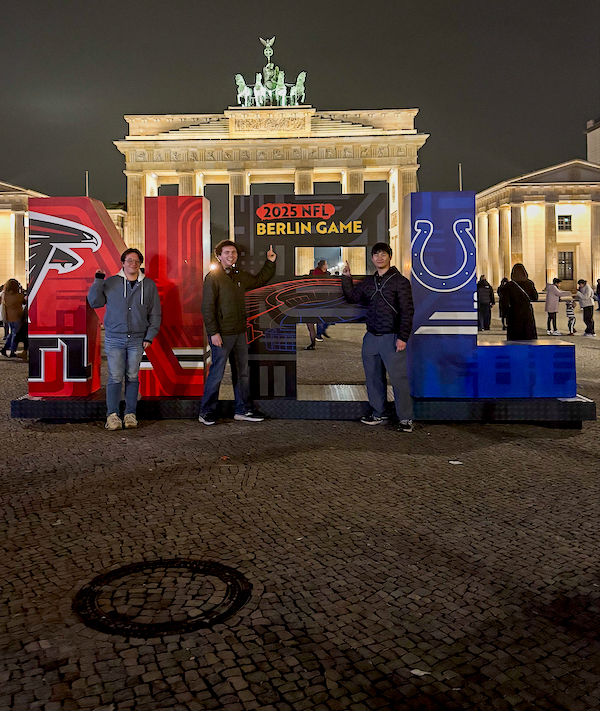 Three people pose in front of illuminated “2025 NFL Berlin Game” signage with team logos, with the Brandenburg Gate lit up in the background at night.