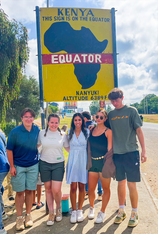 Five people pose on a roadside in front of a large yellow sign reading “Kenya – This Sign Is on the Equator,” with a map of Africa and “Equator, Nanyuki Altitude 6389 ft.” 