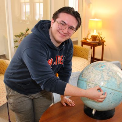 Person in a hoodie leans over a table, pointing at a world globe in a lounge area with chairs and plants.