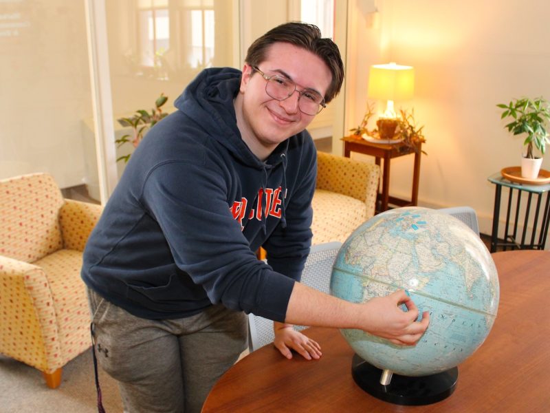Person in a hoodie leans over a table, pointing at a world globe in a lounge area with chairs and plants.