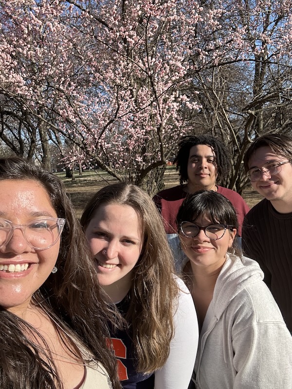 Group of people pose for a selfie outdoors with pink cherry blossoms in bloom behind them. 