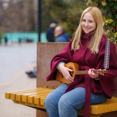 A smiling student sits on a bench outdoors playing a panduri, a traditional Georgian string instrument.
