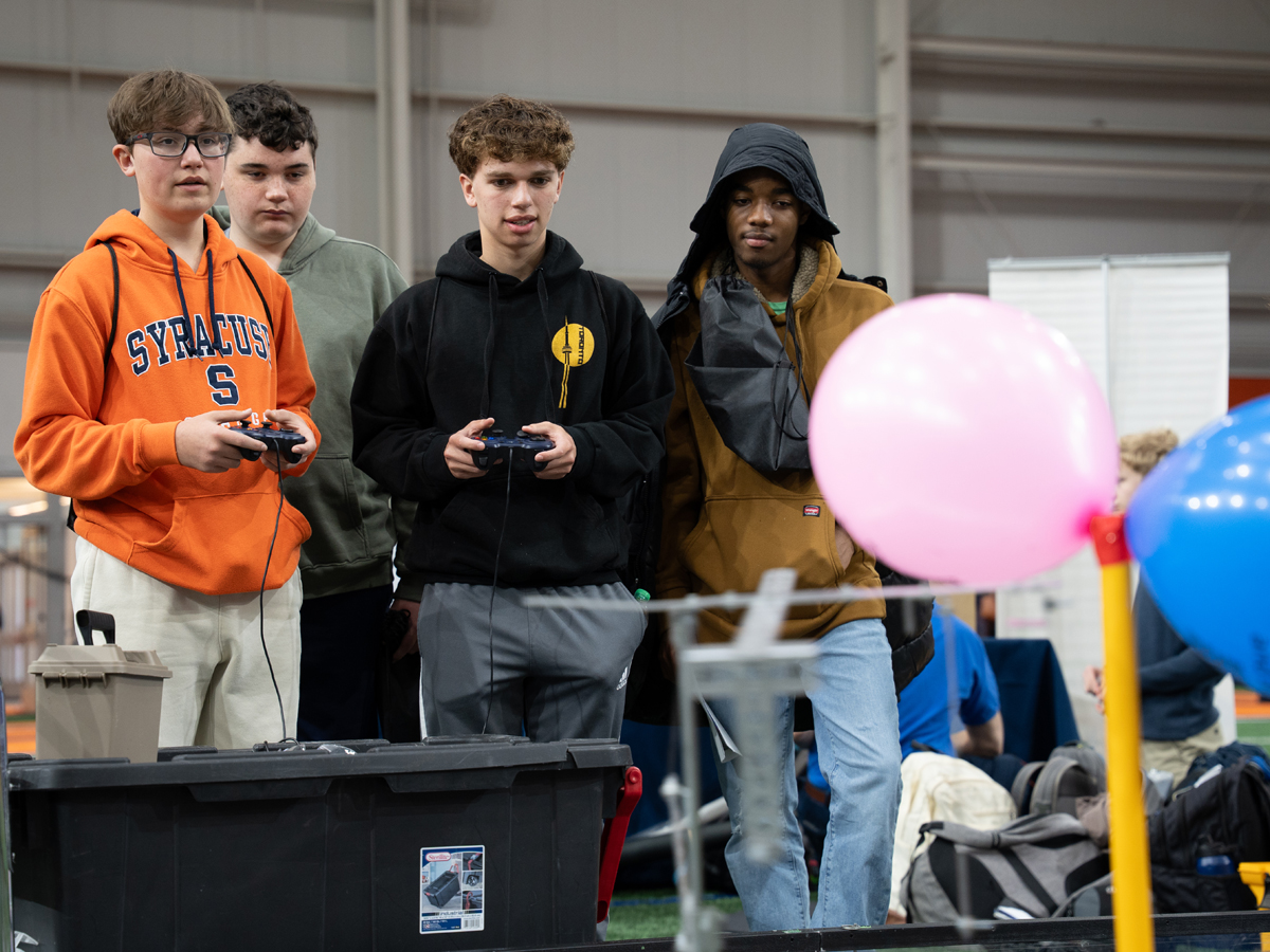 Two students use video game controllers to operate a robot while three students observe.