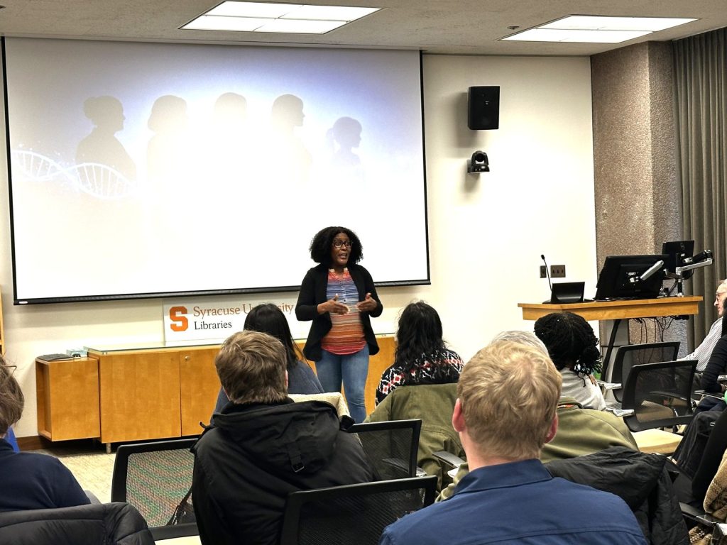 : A 3MT competition contestant speaks to a seated audience with a slide illustration behind her of silhouetted figures. 