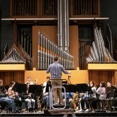 A conductor leads a student orchestra during rehearsal in a concert hall with a large pipe organ in the background.