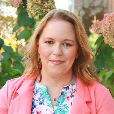 A person photographed outdoors wearing a pink blazer over a floral top, with pink hydrangeas in the background.