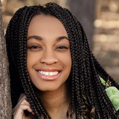 Person with long braided hair stands outdoors beside a tree, facing the camera, with softly blurred foliage in the background.