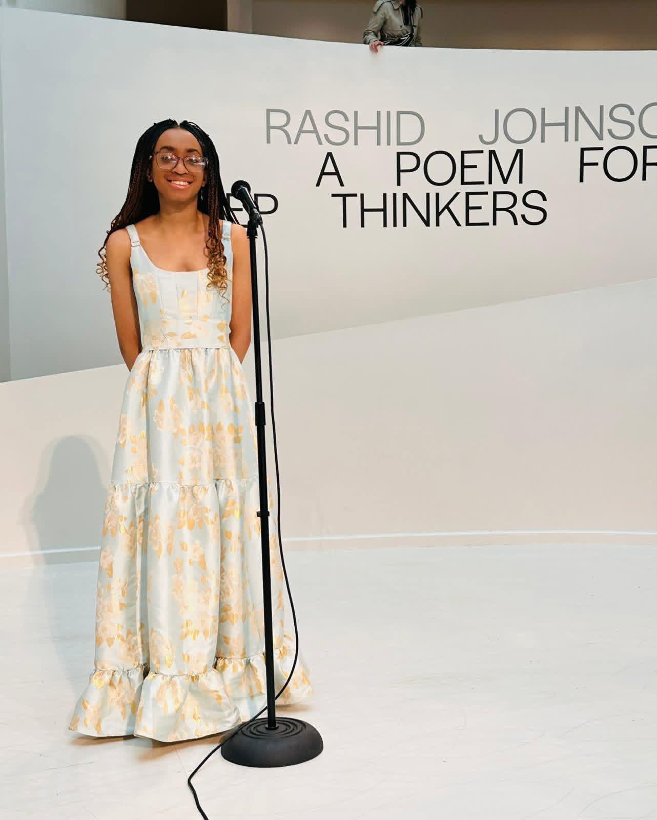 Person stands at a microphone in a gallery space, wearing a long patterned dress, with exhibition text on the wall behind reading “Rashid Johnson: A Poem for Thinkers.”