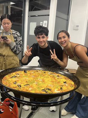 Two students in aprons smile and pose with peace signs behind a large pan of freshly made paella topped with seafood, including mussels and shrimp. A third student photographs the dish in the background.
