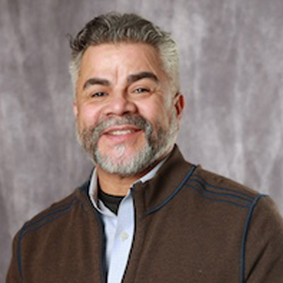 A person smiles in a professional headshot wearing a brown zip-up sweater over a light collared shirt against a gray backdrop.