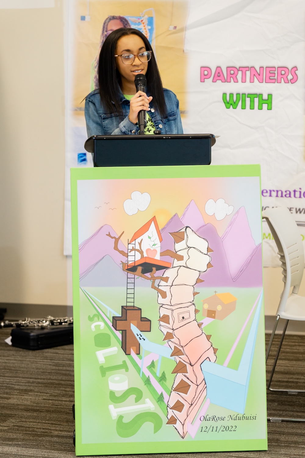 Person speaks into a microphone at a podium, standing behind a colorful illustrated poster, with a branded event banner in the background.
