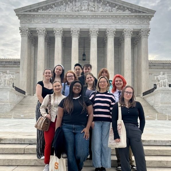 Syracuse University students pose on the steps of the U.S. Supreme Court in Washington, D.C., during the atrocities awareness immersion course. One student holds a Syracuse University tote bag. The court's neoclassical facade and columns are visible behind the group.