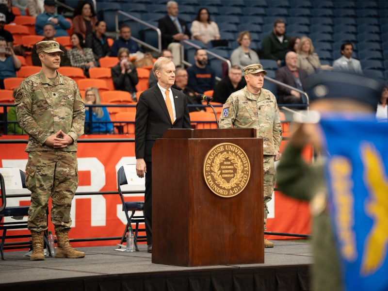 Chancellor Syverud stands at a podium flanked by ROTC officers during a ceremony in a crowded indoor arena
