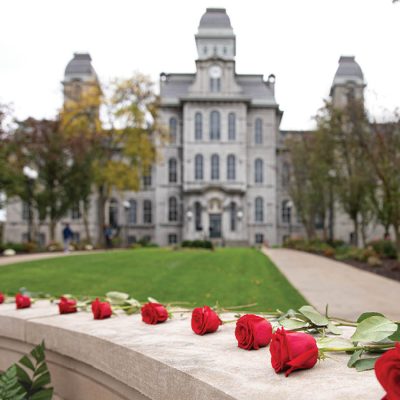Red roses laid along the edge of a stone memorial in the foreground, with Syracuse University's Hall of Languages visible in the soft-focus background on an overcast autumn day.