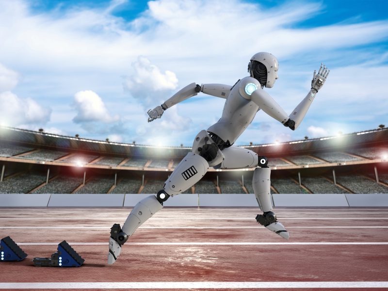 A computer-generated illustration of a white humanoid robot in a running stride on a red track, with starting blocks visible in the background and a stadium crowd under a partly cloudy sky.