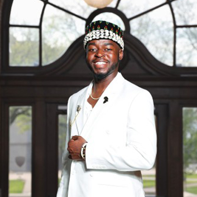 A student smiles in a white suit and beaded cap outside a campus building.
