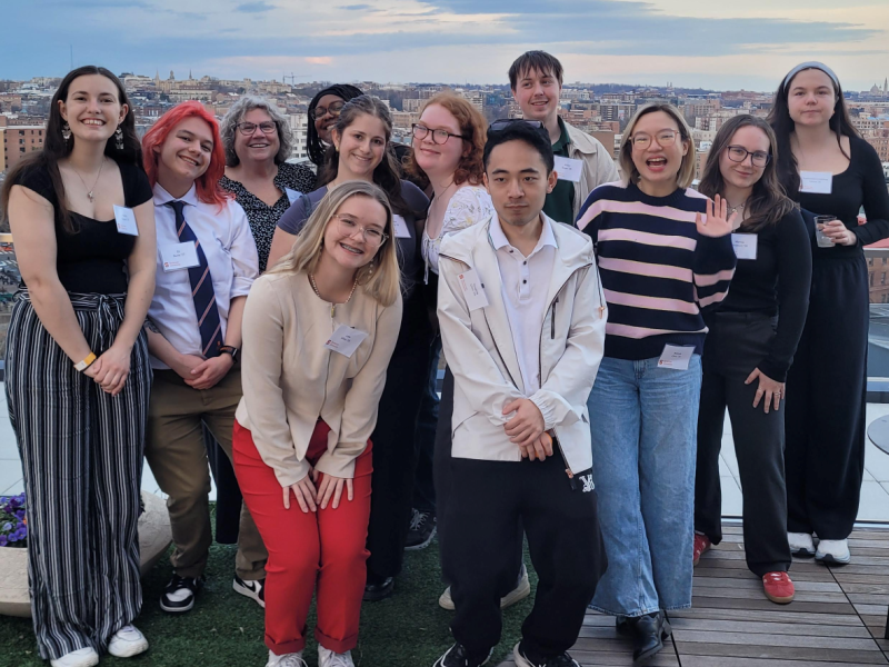 A group of Syracuse University students poses on a rooftop terrace in Washington, D.C., during the atrocities awareness immersion course. The city skyline is visible behind them at dusk. Several students wear name tags and smile for the camera.