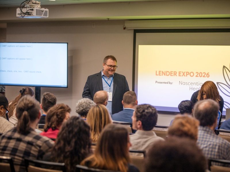 A speaker addresses a full room of attendees at the Lender Expo 2026, presented by Nascentia Health. The speaker stands at the front of the room, smiling, with a projection screen displaying the event title and sponsor behind him and a CART captioning screen visible to the left.