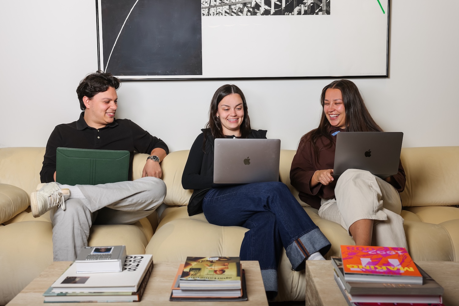Three people seated on a couch, each using a laptop, with books stacked on a coffee table in front of them.