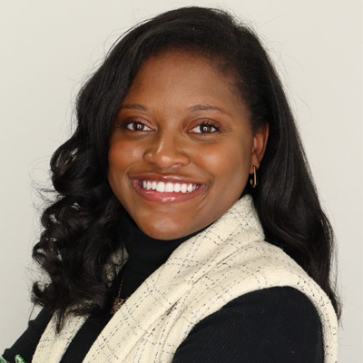 A person smiles in a professional headshot wearing a black turtleneck and cream tweed vest against a light gray background.