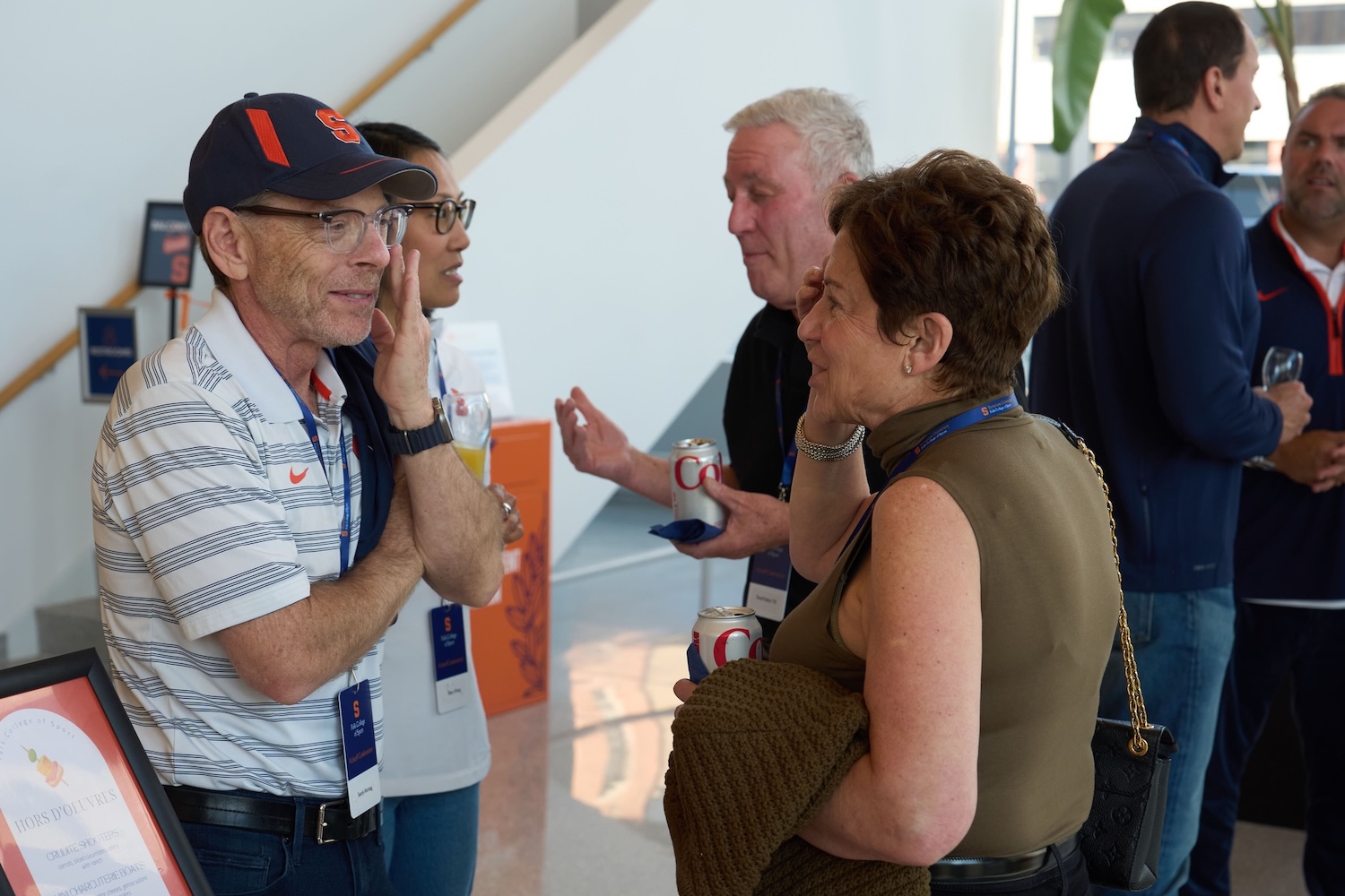 A small group of attendees converse at an indoor event, wearing name badges and holding canned beverages, with other people mingling in the background.