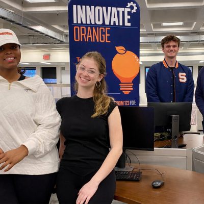 Four students stand in a campus workspace in front of an “Innovate Orange” sign