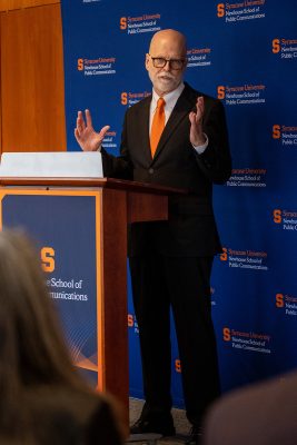 A speaker gestures while delivering remarks at a Syracuse University podium.