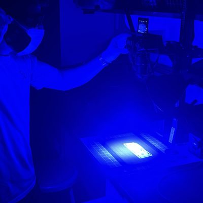 A student in protective gear examines U.S. currency under blue forensic lighting while a camera records.