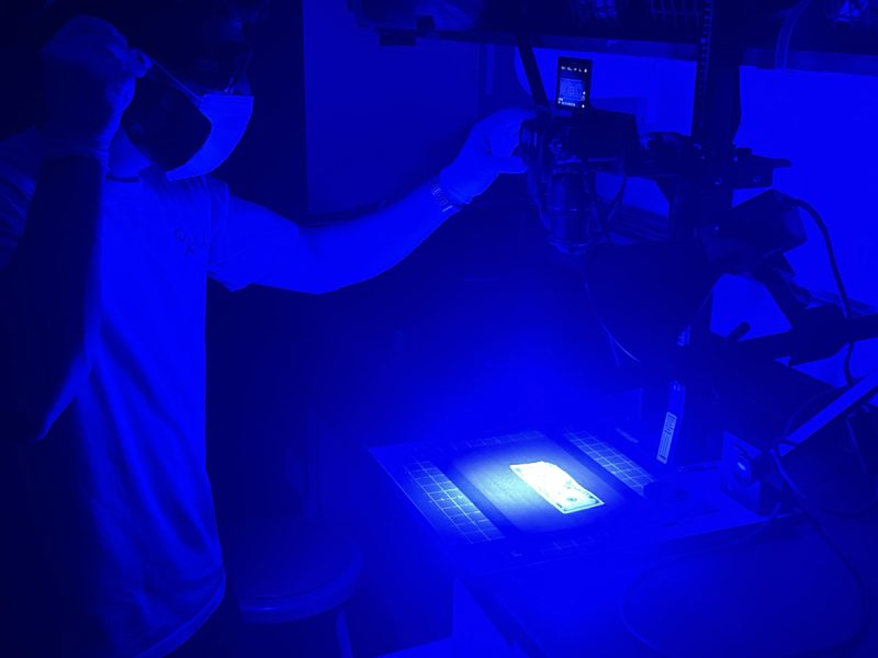 A student in protective gear examines U.S. currency under blue forensic lighting while a camera records.