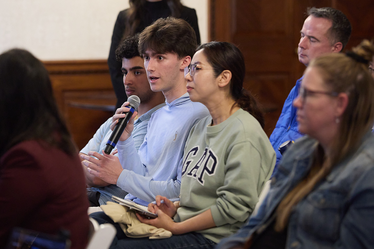 Student holding a microphone and asking a question while seated among peers during a discussion.