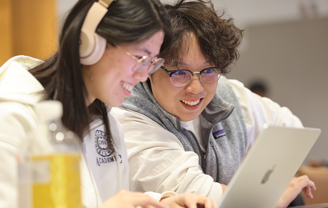 Two students working closely on a laptop at a table, one wearing headphones, with drinks and notebooks nearby. 