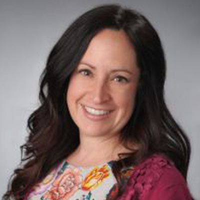 A person smiles in a professional headshot wearing a colorful floral top with a magenta cardigan against a gray background.