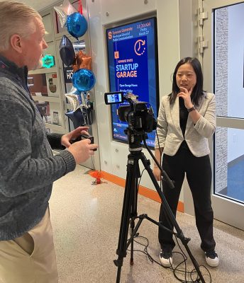 Camera operator films a woman speaking in a hallway decorated with event signage and balloons.