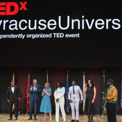 Seven people stand on stage beneath a TEDxSyracuse University banner, flanked by flags, at a past event.
