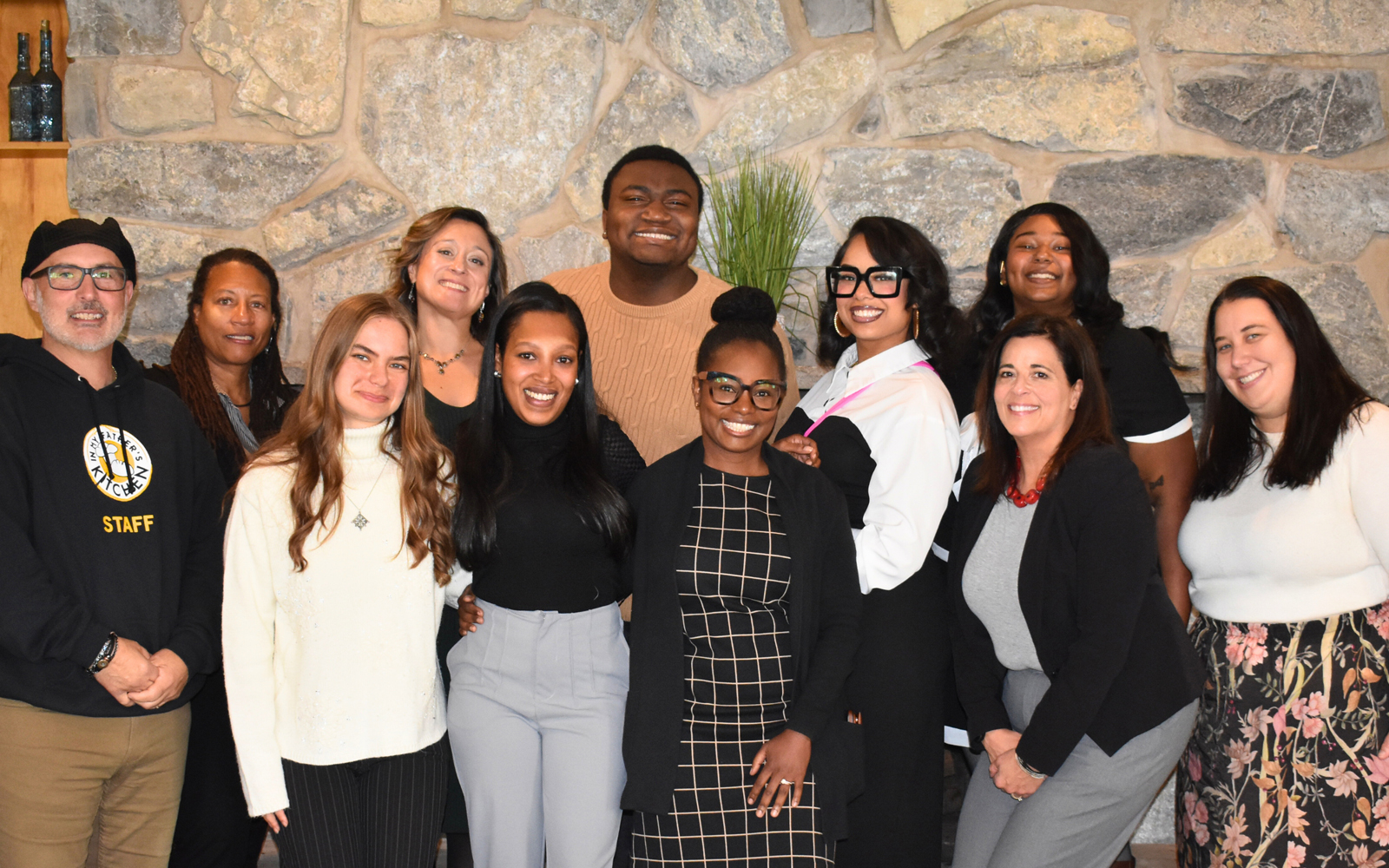 A group of 11 people smile together in front of a stone wall at an indoor gathering.