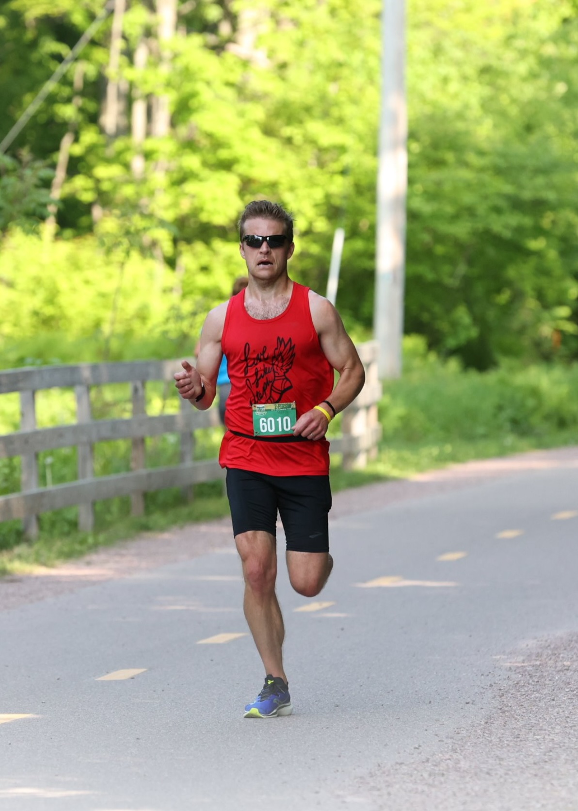 Runner wearing a red race singlet and bib number 6010 runs on a paved trail bordered by a wooden fence and trees.