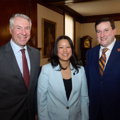 Three professionals smiling and standing together in a formal meeting room with framed portraits on the wall