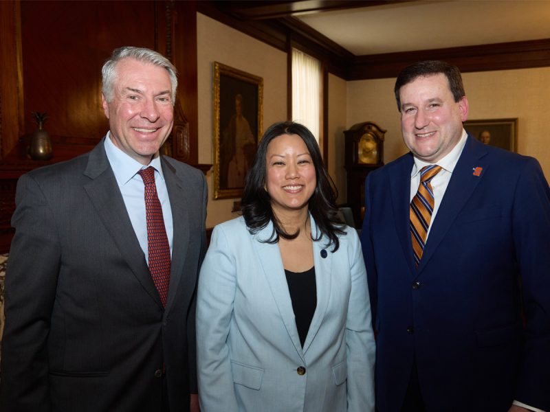 Three professionals smiling and standing together in a formal meeting room with framed portraits on the wall