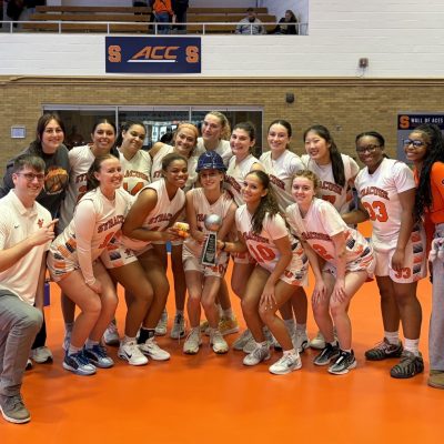 Basketball team in orange‑and‑white jerseys poses with a trophy on an indoor court, with an American flag and ACC signage in the background.