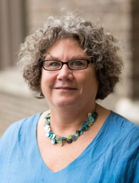 A person with curly chin length grey hair and glasses wearing a blue top and a blue and green necklace poses outdoors.