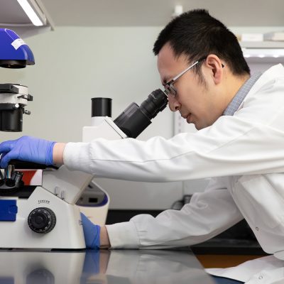 A professor in a white lab coat and blue gloves looks through a microscope.
