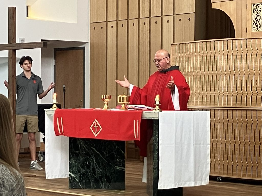 A Catholic priest in red vestments celebrates Mass at a decorated altar, with a young altar server standing near a wooden cross in the background.