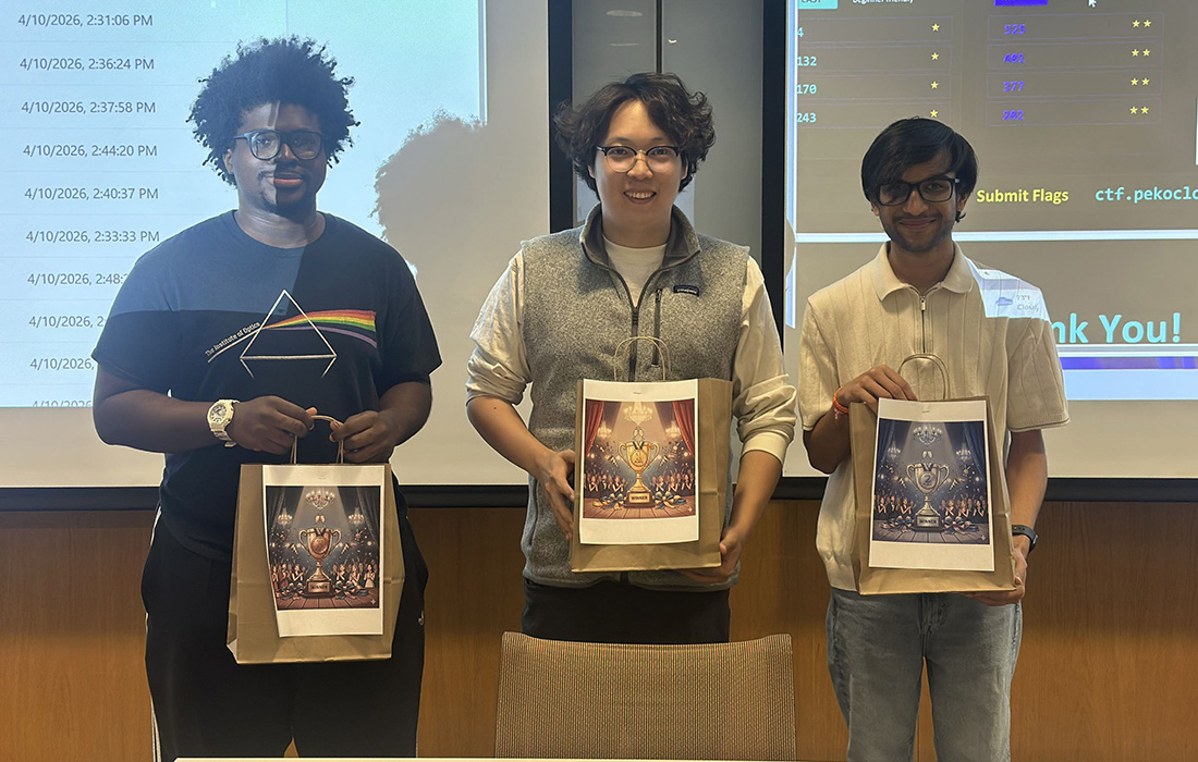 Three people standing in a classroom holding gift bags, with a presentation screen visible behind them.